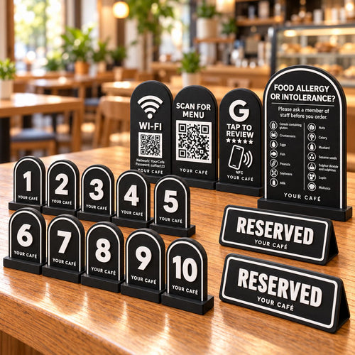 Café Starter Kit in black: ten numbered table signs, Wi-Fi QR sign, menu QR stand, NFC review card, allergen sign and two reserved signs on a café table