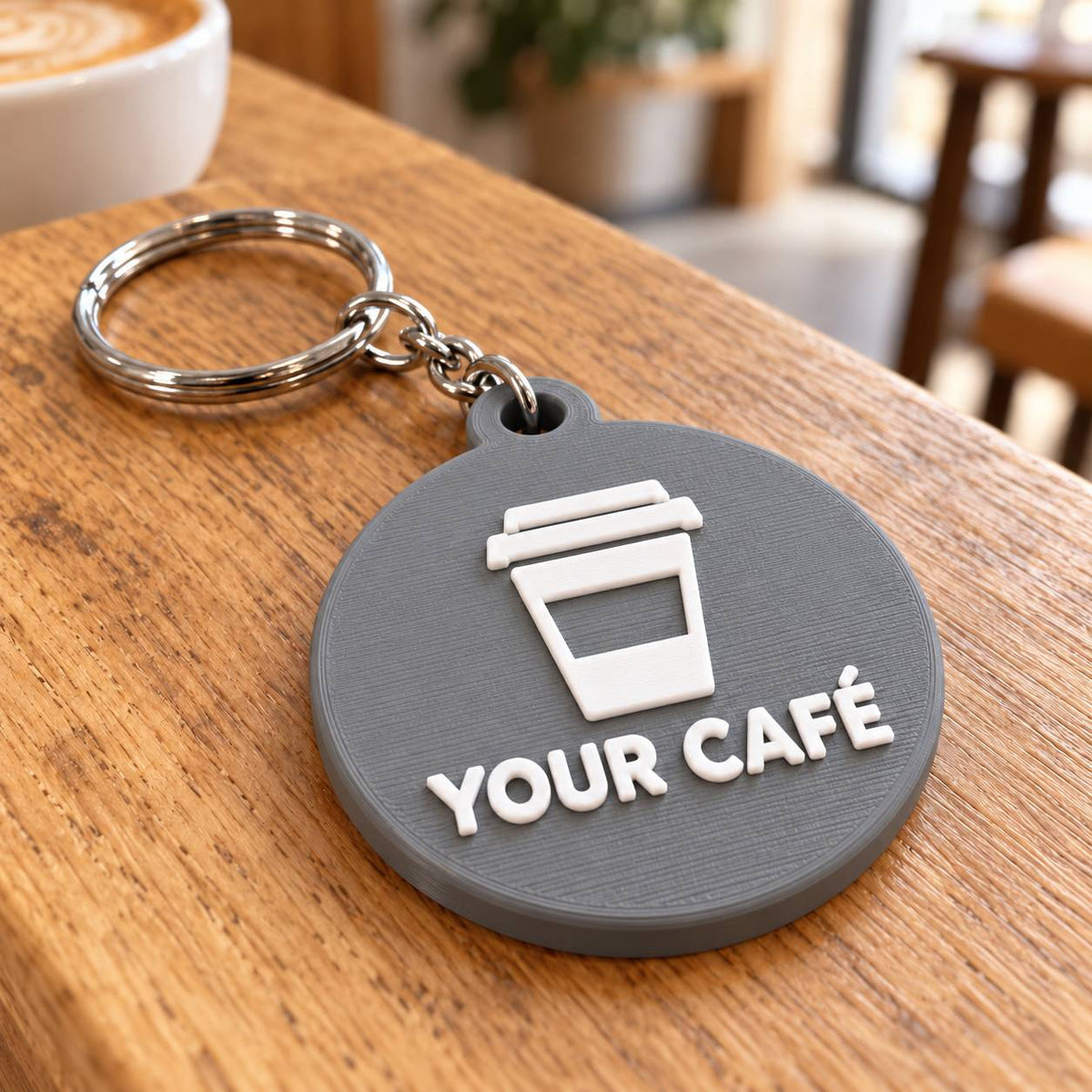 Round grey 3D printed keyring with takeaway coffee cup logo and café name on a wooden table